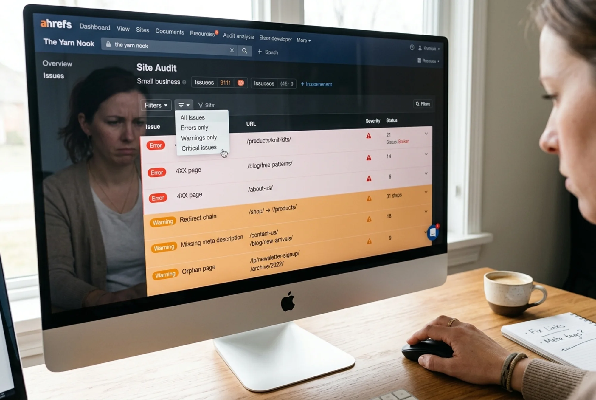 A monitor filling the frame with the Ahrefs Site Audit issues panel — long stacked rows of red "Error" and orange "Warning" rows labeled "4XX page," "Redirect chain," "Missing meta description," "Orphan page" — and a small business owner's hand hovering with the cursor over a filter dropdown, looking unsure where to start