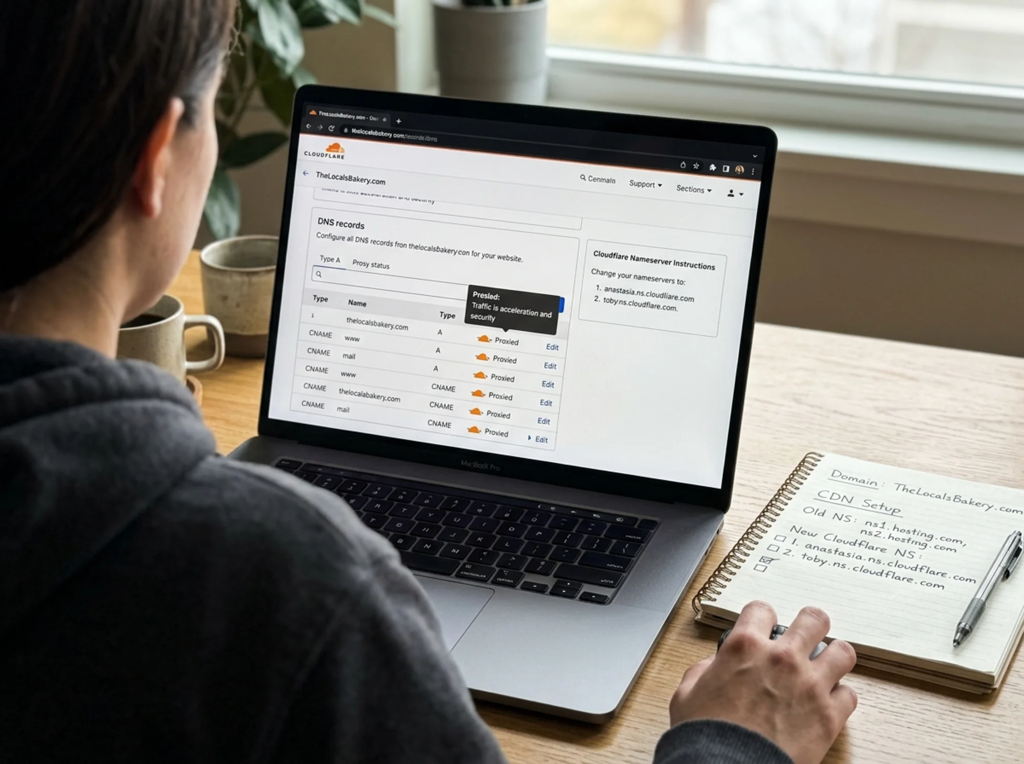 A close-up over-the-shoulder view of a Cloudflare dashboard mid-setup, DNS records being toggled to "Proxied" status with orange cloud icons, nameserver instructions visible, a notebook beside the laptop with handwritten domain notes