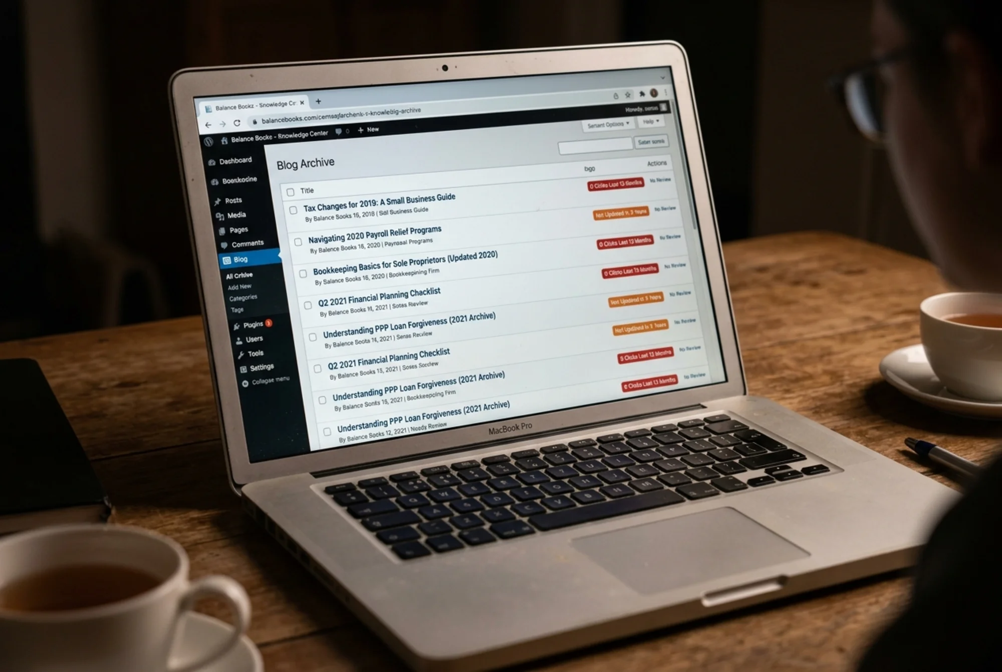 Laptop screen showing a sprawling blog archive page for a small bookkeeping firm, with post titles dated 2019-2021, visible labels like "0 clicks" and "not updated in 3 years" next to each entry, dim lighting suggesting neglect