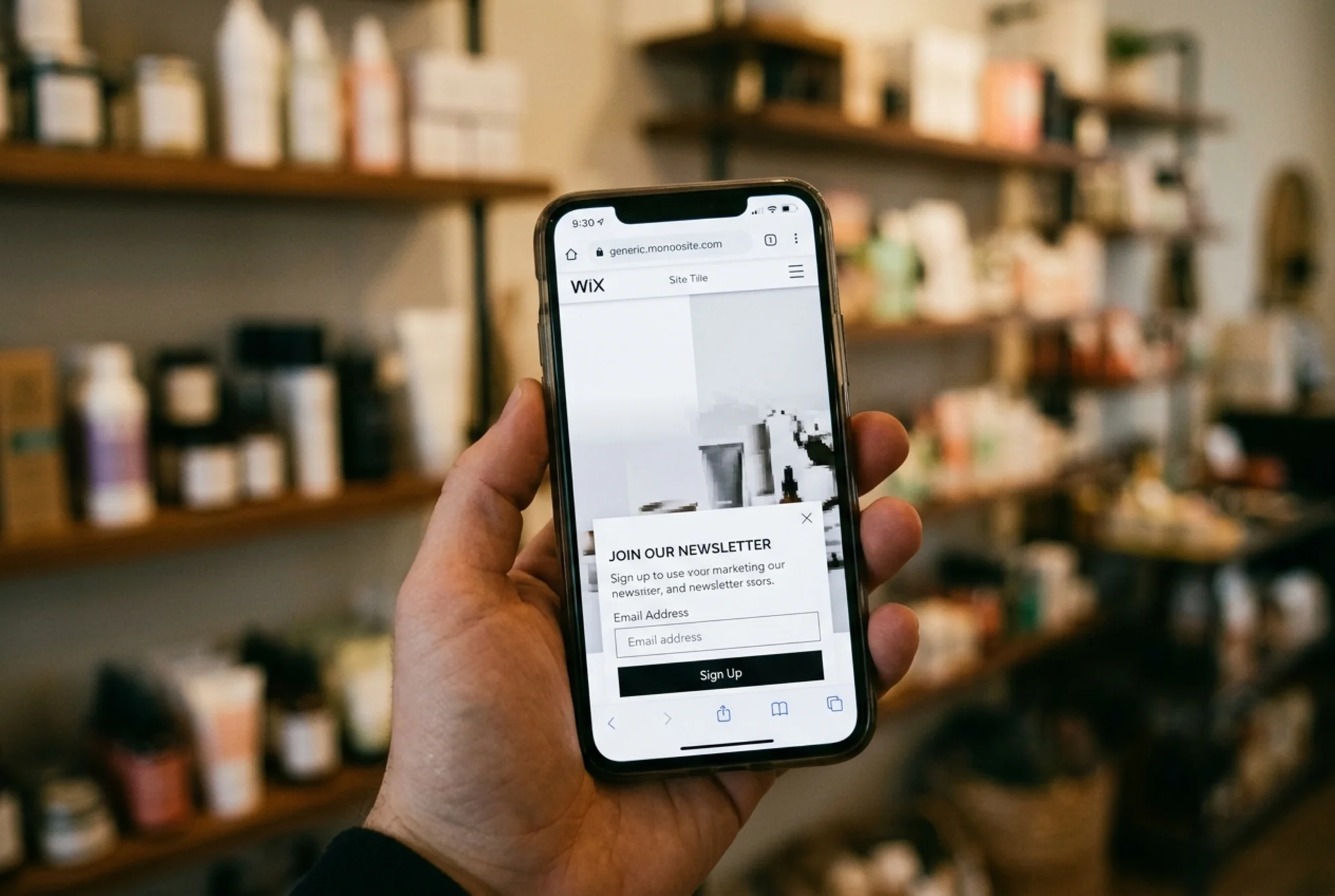 Close-up of a mobile phone showing a partially loaded Wix homepage with a massive hero image still rendering, a delayed newsletter popup half-dropped from the top, blurred boutique shelves in the background