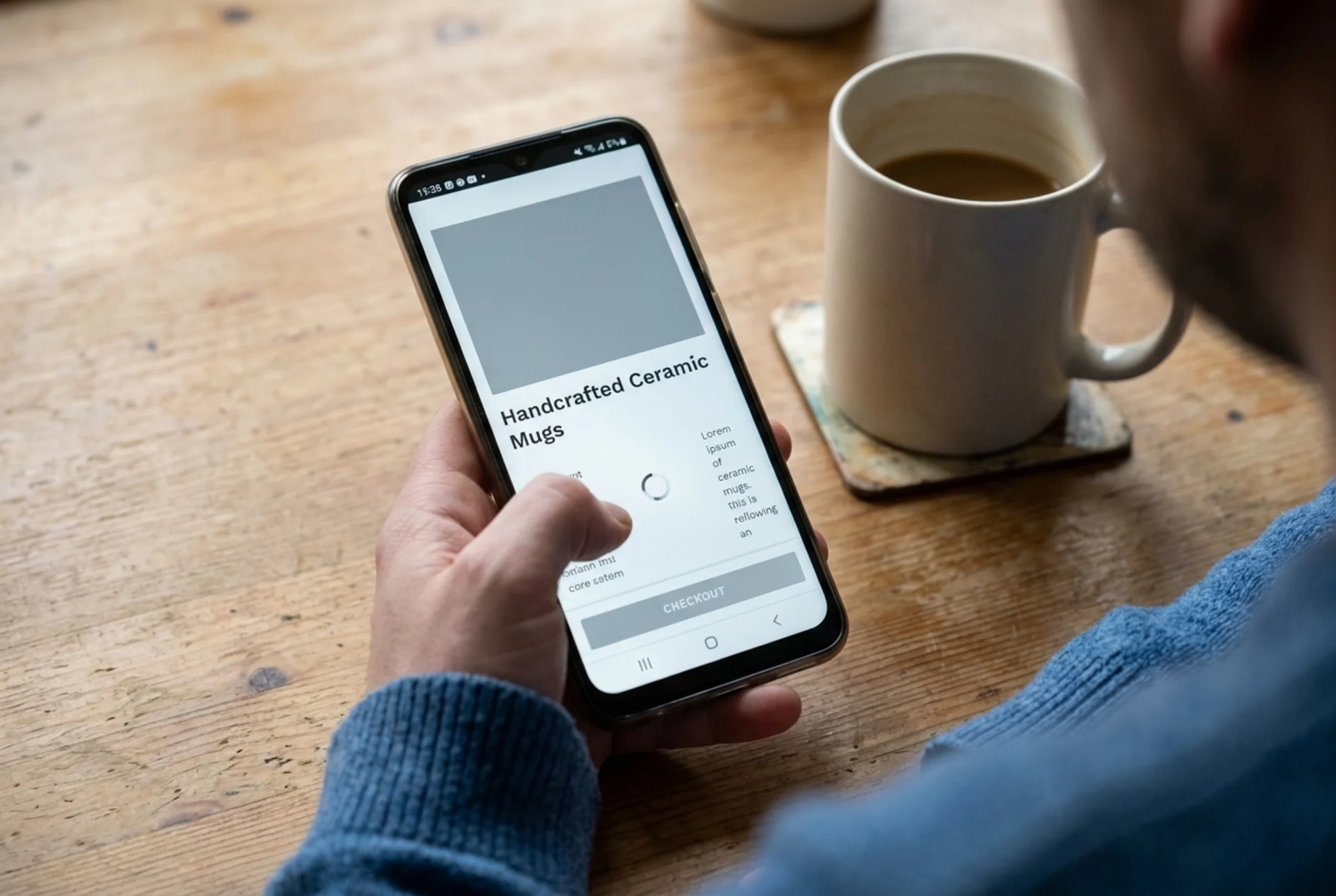 Mid-range Android smartphone on a wooden kitchen table showing a half-loaded ceramic mug product page, large grey placeholder where the hero image should be, spinning loader, reflowing text, a stalled checkout button, a frustrated shopper's thumb hovering near the back arrow
