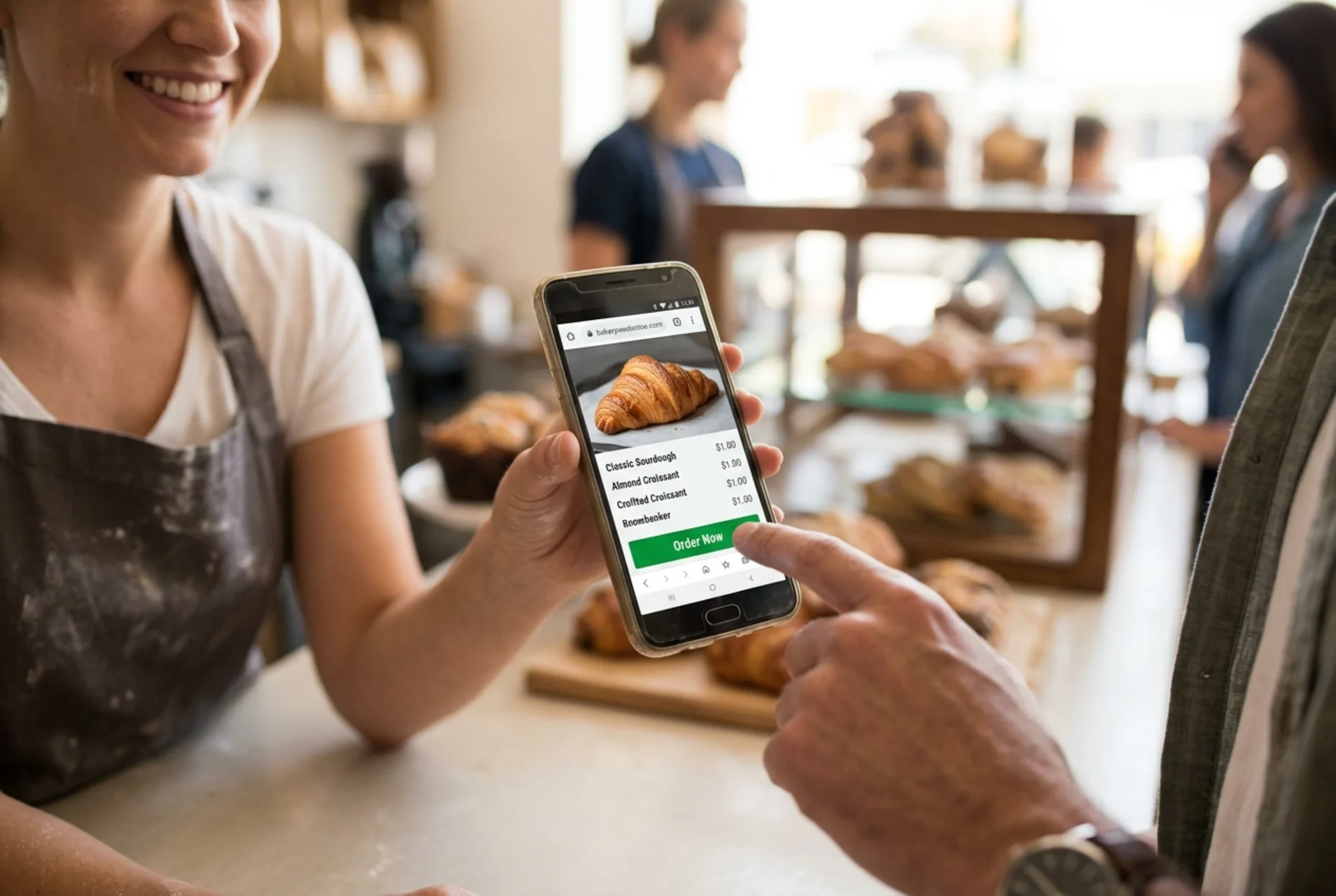 Same budget Android phone now showing the bakery homepage fully rendered with a crisp croissant photo, readable menu, and a green "Order Now" button visible above the fold, owner smiling as a customer's finger taps the button