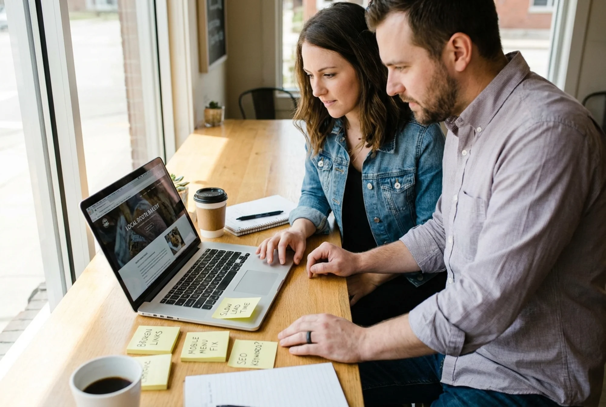 A small business owner and a web consultant sitting together at a table, looking at a laptop screen showing a website with sticky notes on the table listing audit findings